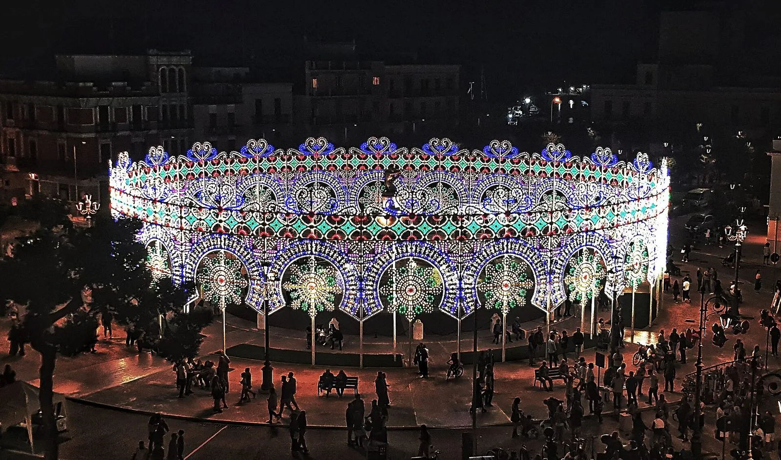 Una grande luminaria circolare ad archi colorati accesa di notte in una piazza di Mola di Bari durante la festa patronale, con persone che passeggiano