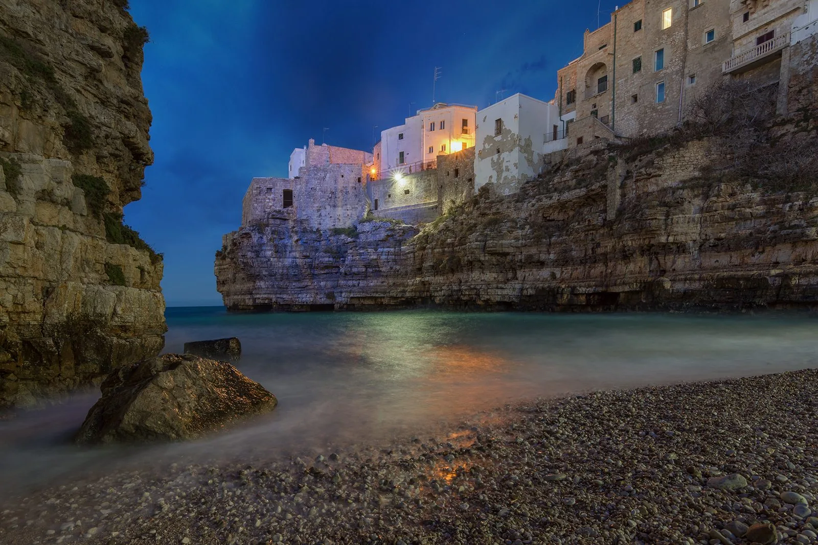 La spiaggia di ciottoli della Lama Monachile all'ora blu, con le case illuminate del centro storico di Polignano a Mare sulla scogliera